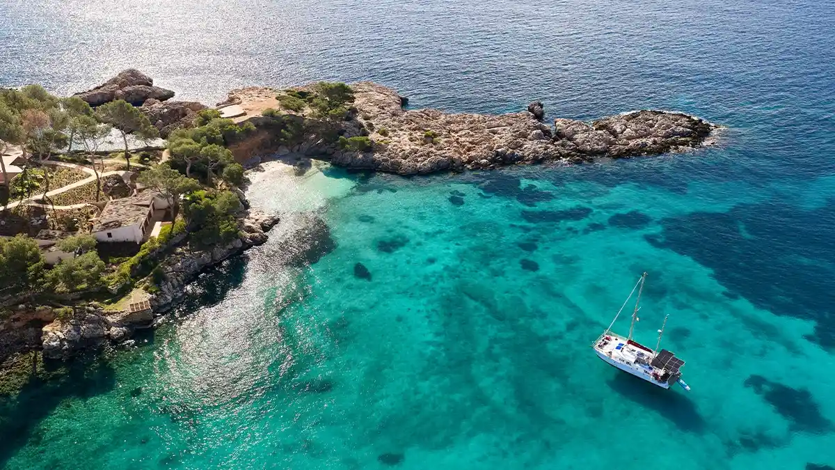 Aerial view of a yacht anchored off a rocky pine-covered headland in clear turquoise water