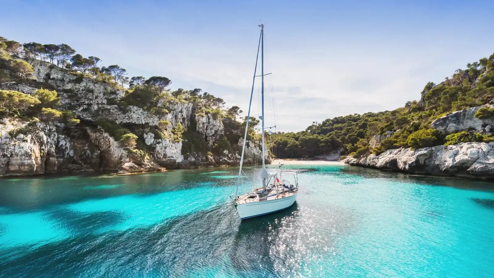 A sailing yacht at anchor in a turquoise cove between pine-topped limestone cliffs