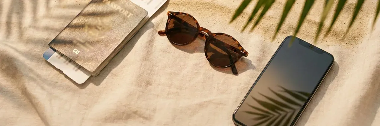 A passport with boarding pass, sunglasses and a phone on a sunlit beach blanket under palm shadows